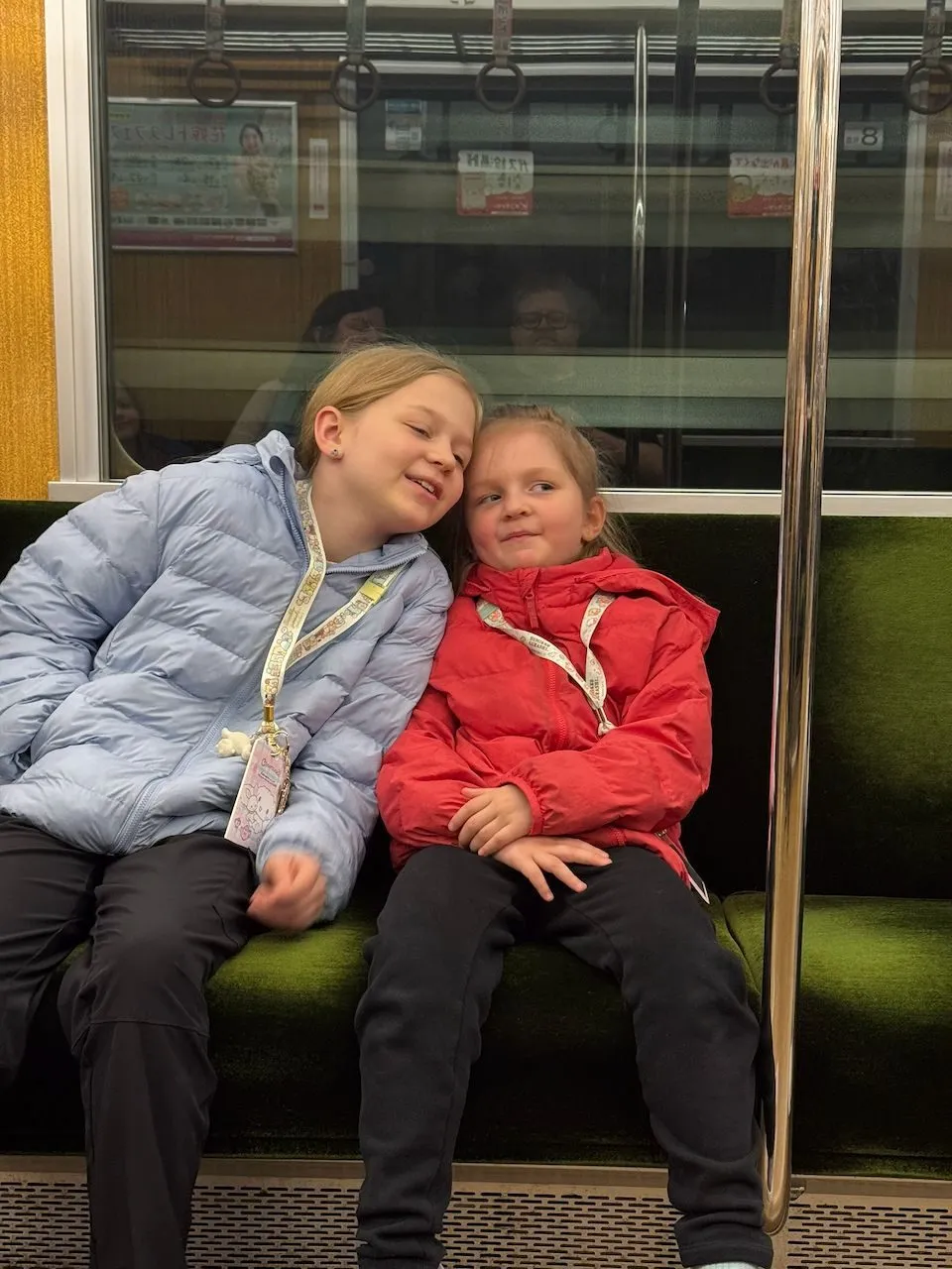 Two girls sitting together on a green-seated Osaka subway car, the older one leaning her head toward the younger one, both wearing Sanrio lanyards and winter jackets.