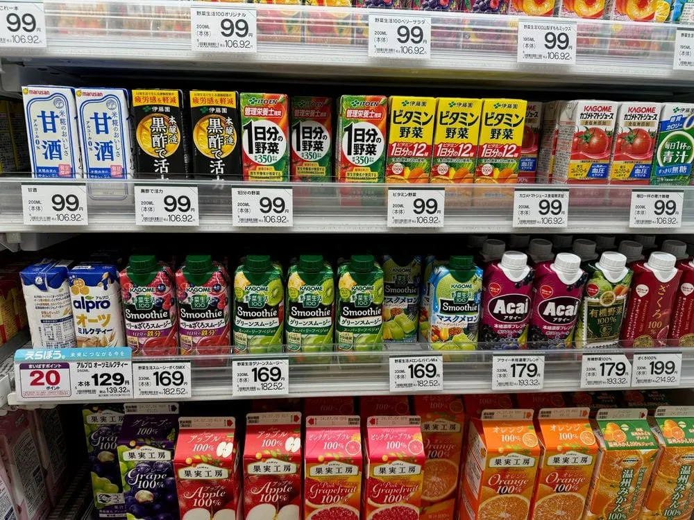 A cold-drink shelf at a Japanese supermarket in Shibamata, Tokyo, with rows of juice, smoothies, and amazake bottles under yellow price tags reading 99, 169, 179, and 199 yen.