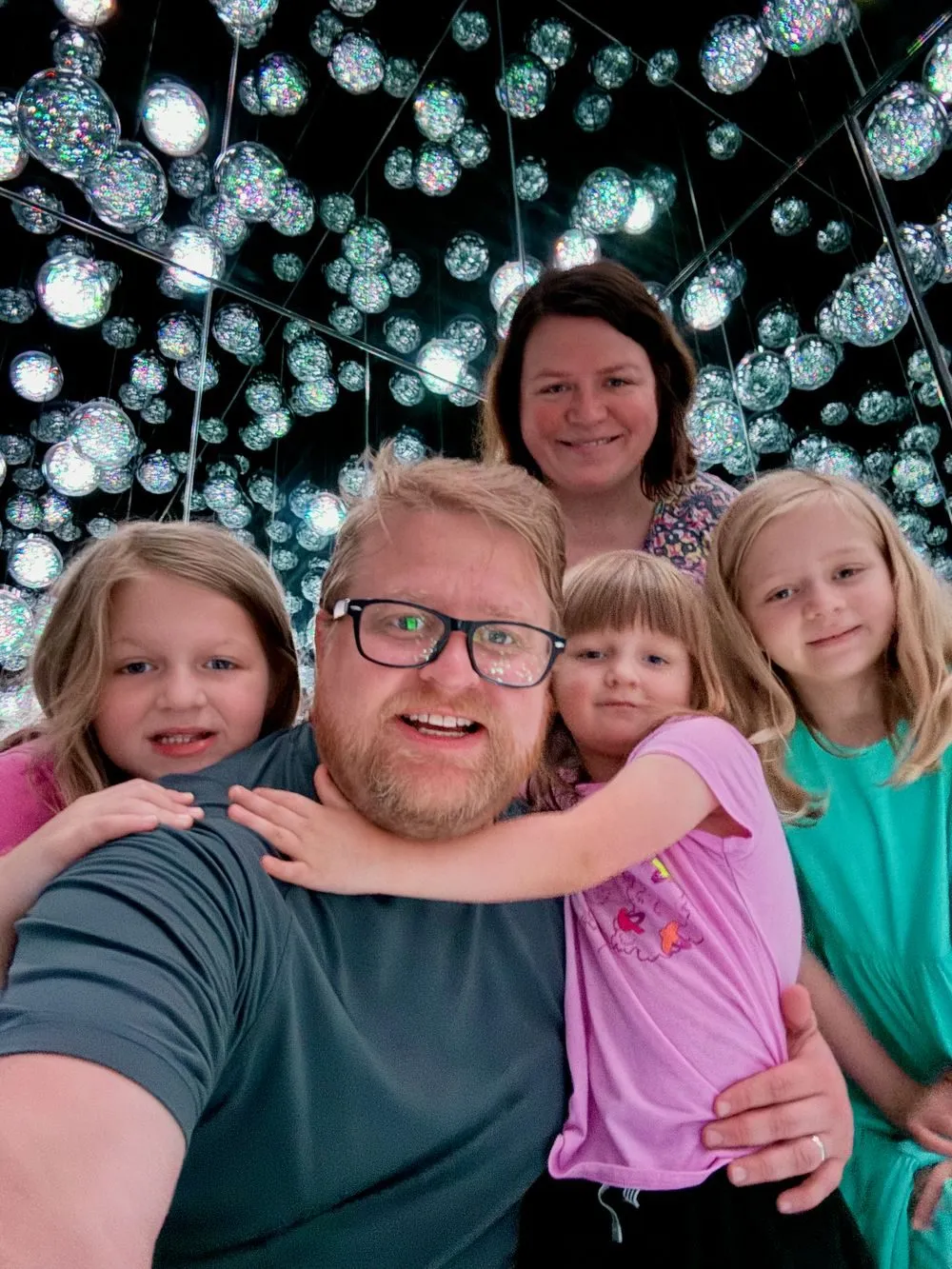 A family of five takes a selfie inside a dark room filled with hundreds of suspended iridescent light orbs at teamLab Borderless in Tokyo, with the father in the foreground holding a young child and two older girls and a woman smiling behind him.