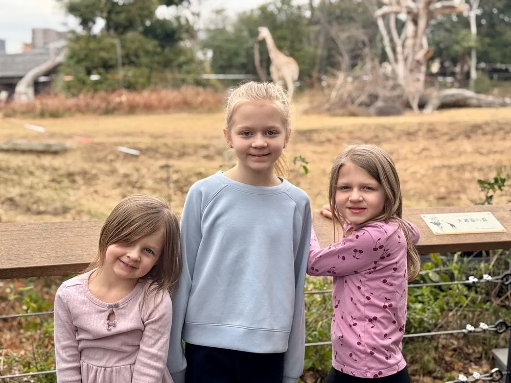 Three girls smile at the camera in front of a railed enclosure at Tennoji Zoo in Osaka, with a giraffe visible in the background photobombing the shot.