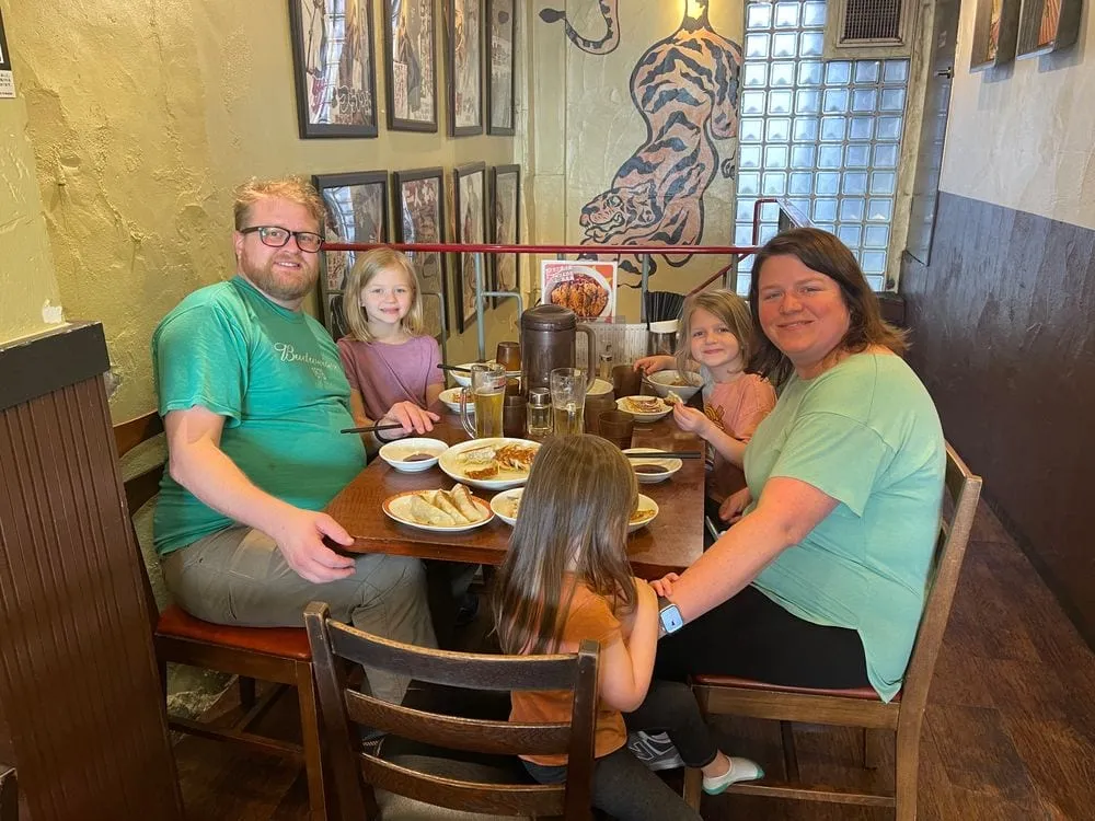 A family sits at a wooden counter inside a retro gyoza restaurant in Asakusa, Tokyo, with a painted tiger mural and framed photos on the wall behind them.
