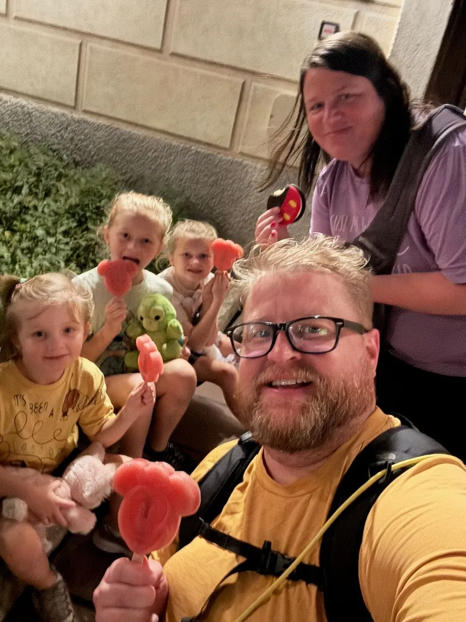 A man takes a selfie holding a Mickey Mouse-shaped ice cream bar in the foreground, while a woman and three young girls seated behind him each hold up their own Mickey-shaped treats outside at night near a stone wall.