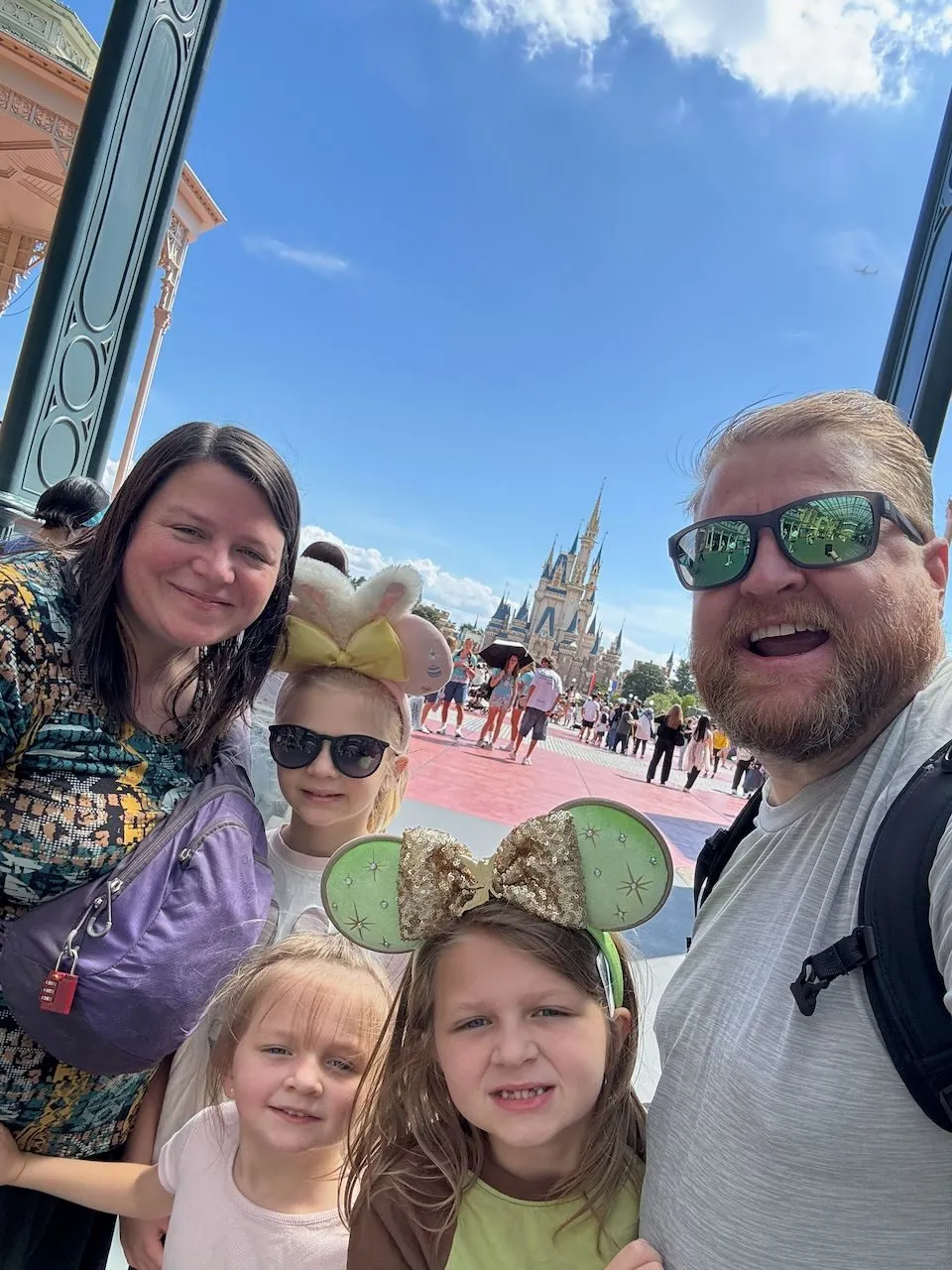 A family of five takes a selfie in front of Cinderella's Castle at Tokyo Disneyland, with two adults and three young girls, one wearing Minnie Mouse ears with a gold sequin bow and another with a stuffed character headband and sunglasses.