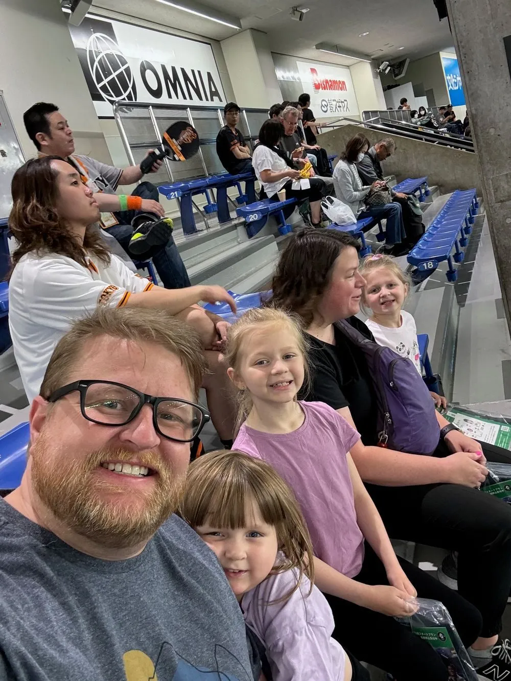 A family of five takes a selfie in stadium seats at Tokyo Dome before an evening baseball game, with other fans visible in the rows behind them.