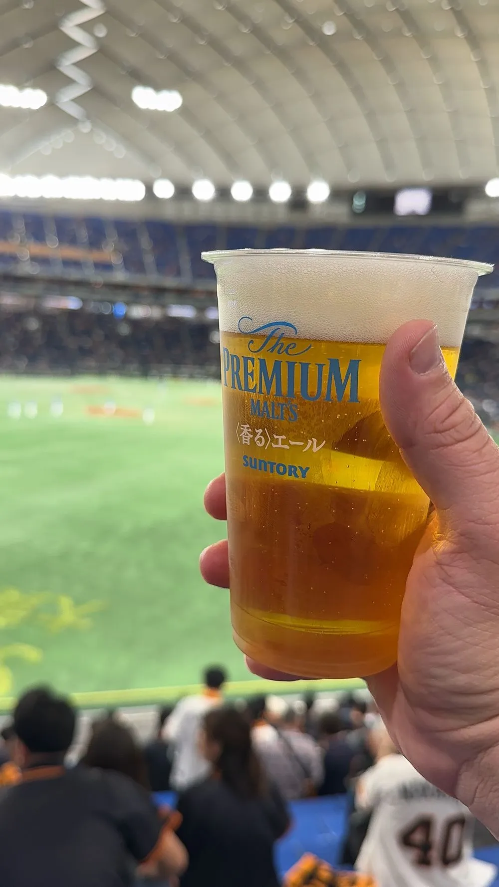 A hand holds a plastic stadium cup of draft beer branded The Premium Malt's, with a baseball field visible in the background at Tokyo Dome.