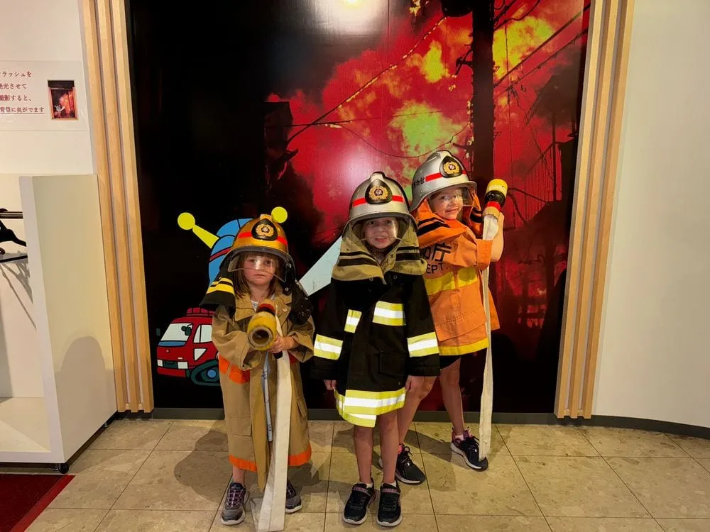 Three young children wearing yellow and orange firefighter costumes and helmets stand in front of a Tokyo Fire Department exhibit display.