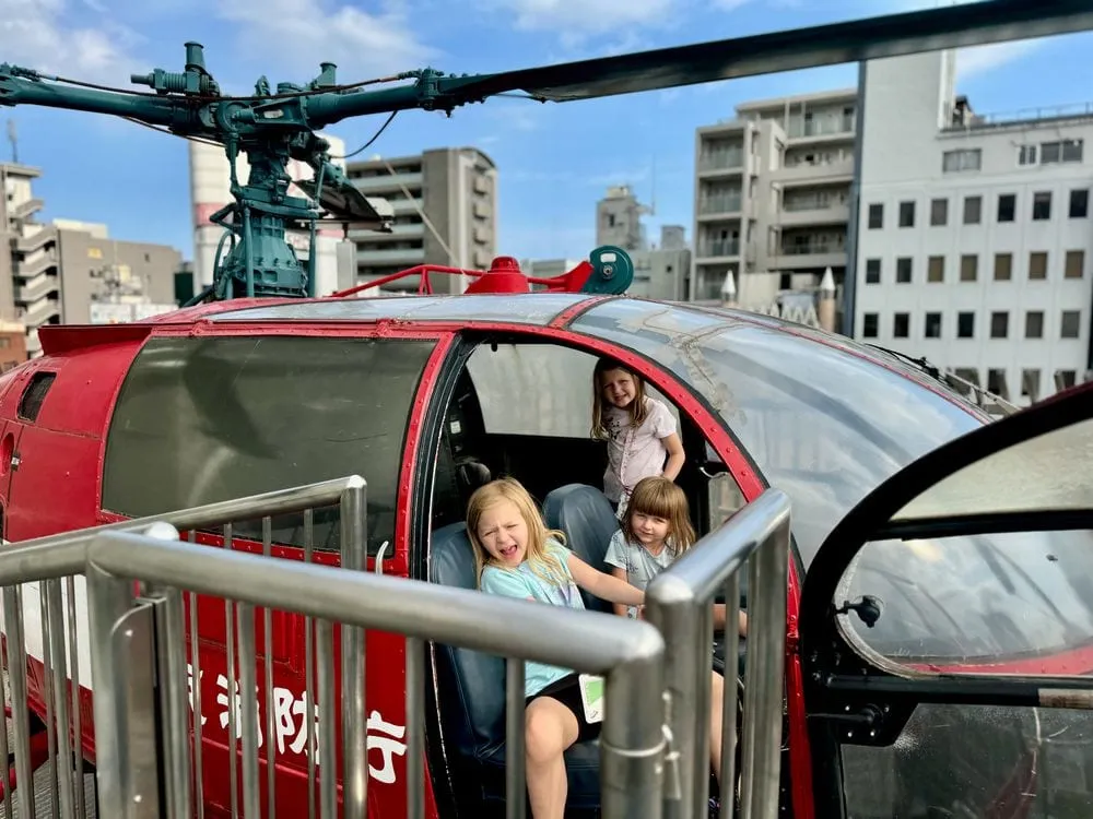 Three young girls sit inside a red fire department helicopter cockpit on the rooftop of the Tokyo Fire Museum in Yotsuya, Shinjuku.