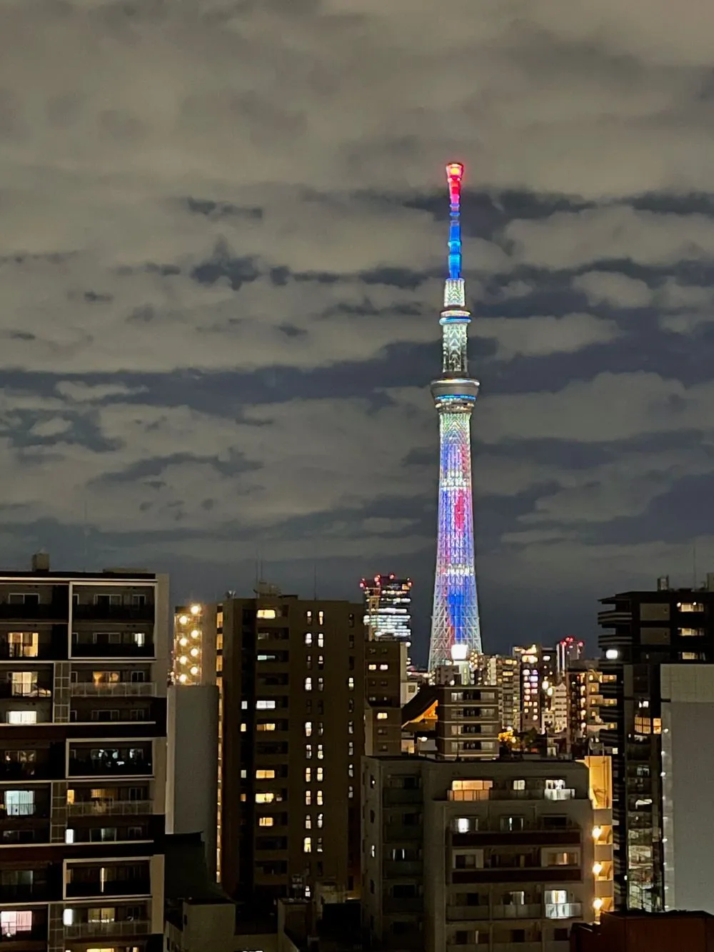 Tokyo Skytree lit up at night in a colorful illumination pattern, rising above a dense cityscape of illuminated apartment buildings.