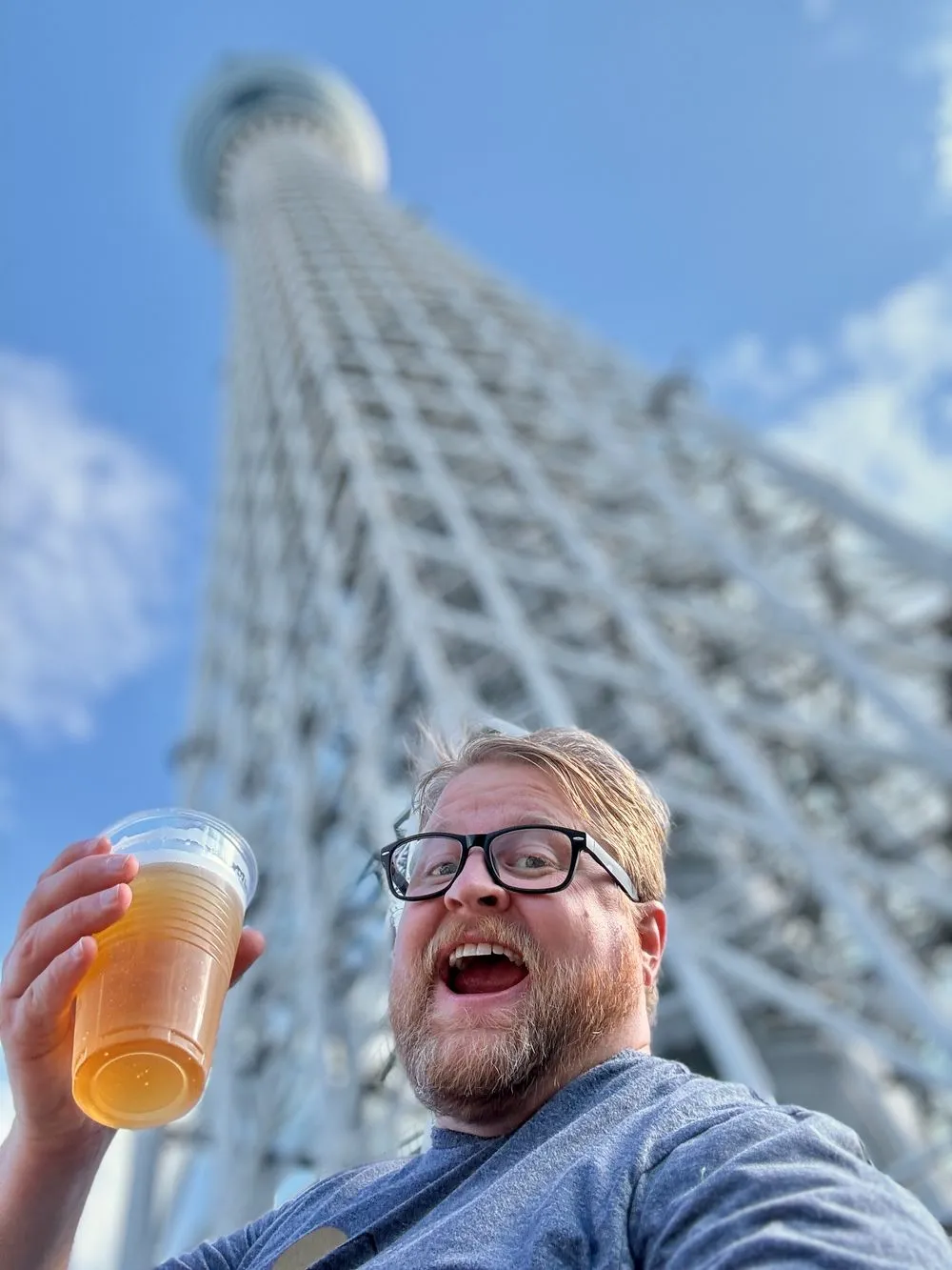 A man wearing glasses holds up a plastic cup of beer with Tokyo Skytree towering directly behind him against a blue sky.
