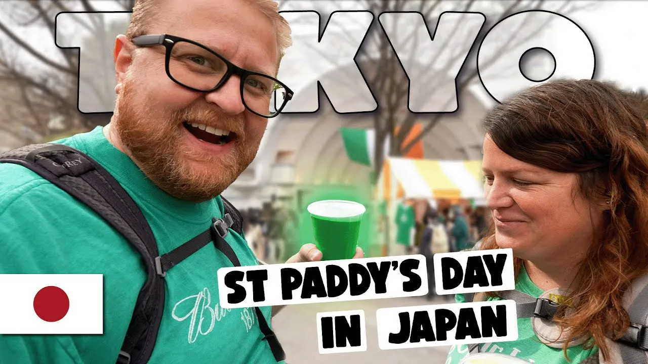 A YouTube thumbnail of Adam and Lindsay at Tokyo's St. Patrick's Day festival, Adam holding a green beer, green bunting visible at the festival stalls behind them.