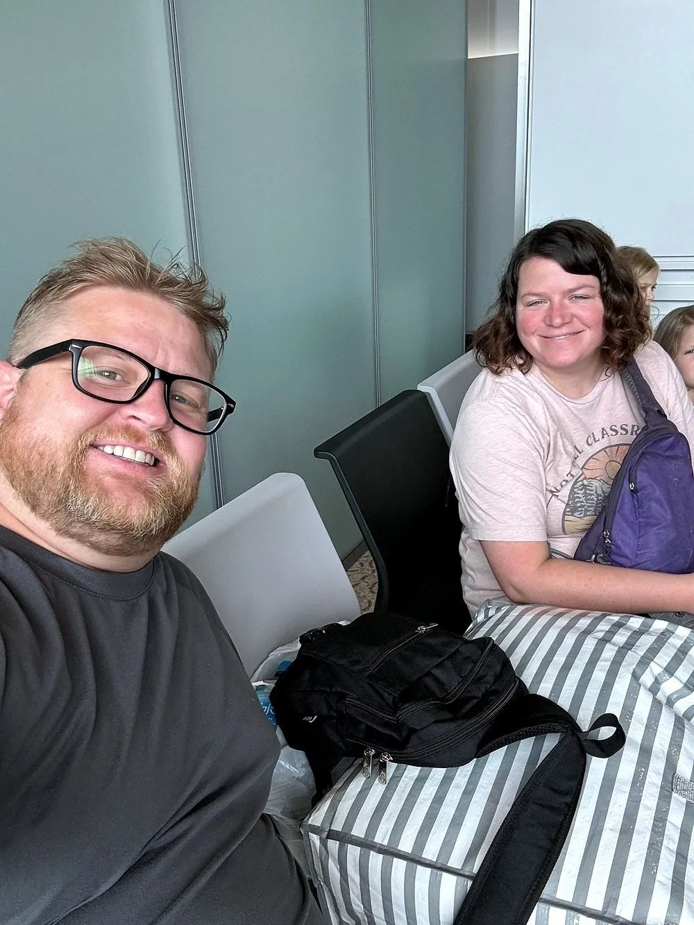 A man with glasses and a beard takes a selfie at an airport gate while a smiling woman in a light pink t-shirt sits behind him next to two children, with luggage stacked on the seats in front of them.