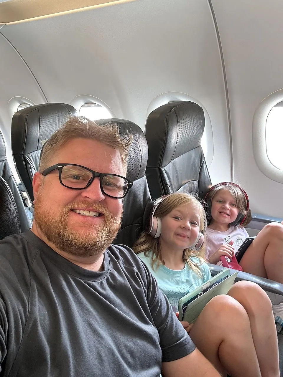 Adam takes a selfie at Narita Airport with Lindsay smiling behind him, bags stacked on the seat between them, a child partially visible beside Lindsay.