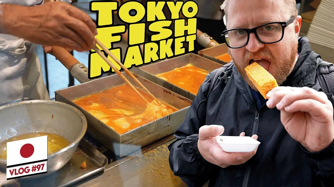 A YouTube thumbnail of Adam eating a skewer of tamagoyaki at Tsukiji Outer Market in Tokyo, with vendors preparing food behind him.