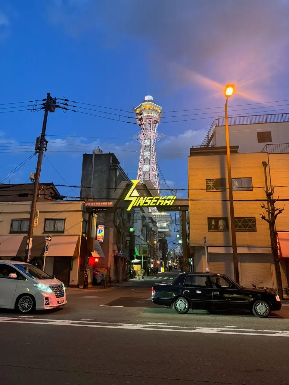 A street-level view at blue hour of the illuminated Tsutenkaku tower rising above the Shinsekai district entrance gate in Osaka, with a black taxi and white van in the foreground.