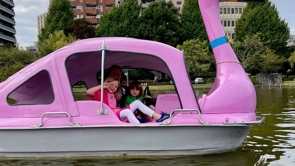 Two young girls ride together in a pink swan-shaped paddle boat on Shinobazu Pond in Ueno Park, Tokyo, with city buildings visible across the water.