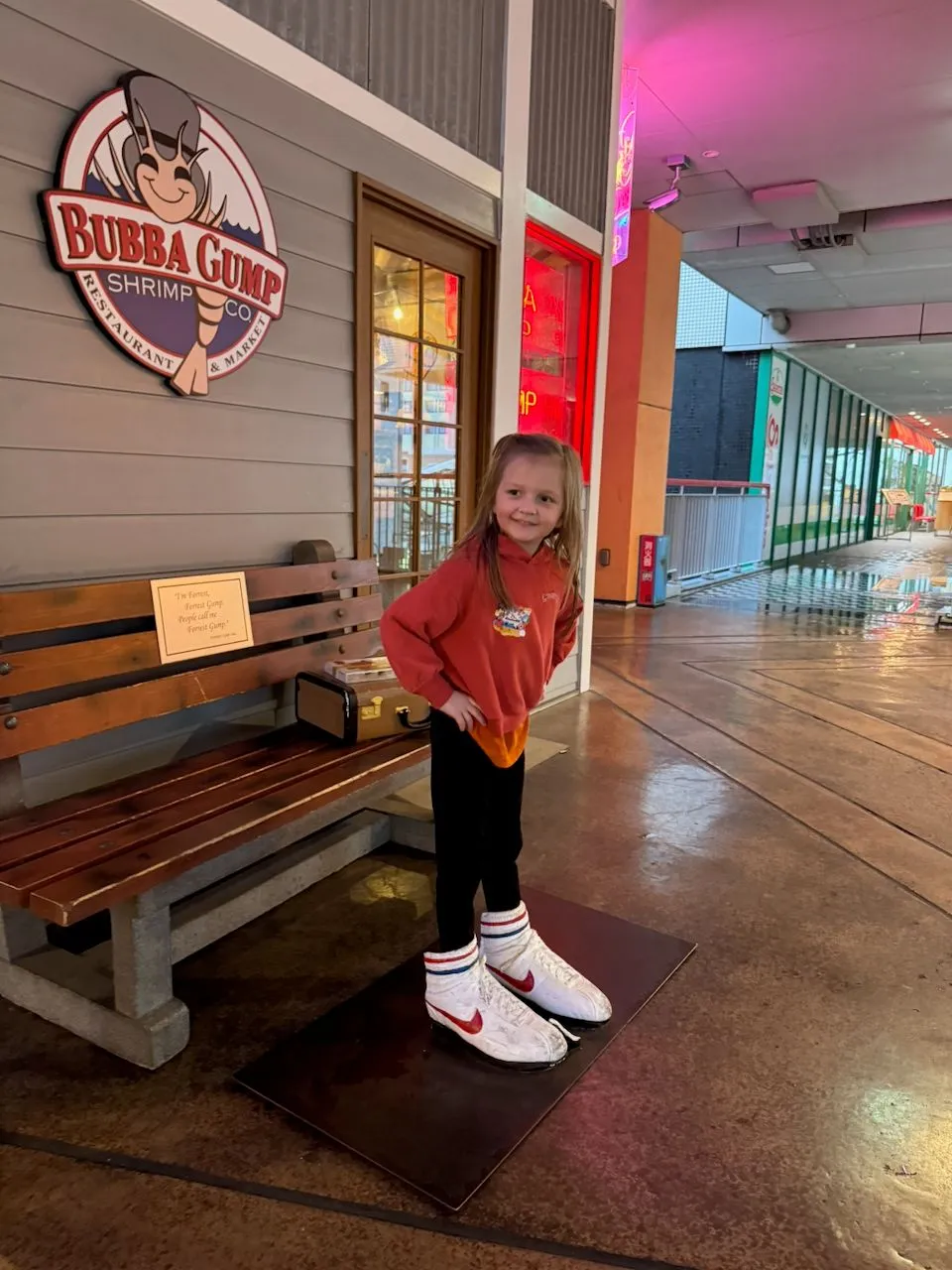 A young girl in an orange hoodie stands smiling on a pair of oversized Forrest Gump shoe props outside Bubba Gump Shrimp Co. at Universal CityWalk Osaka.