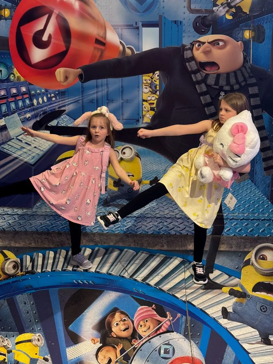 Two young girls pose in front of a large Despicable Me interactive photo backdrop featuring Gru and Minions, one in a pink dress with bunny ears and one in a yellow dress holding a Hello Kitty plush, both striking action poses.