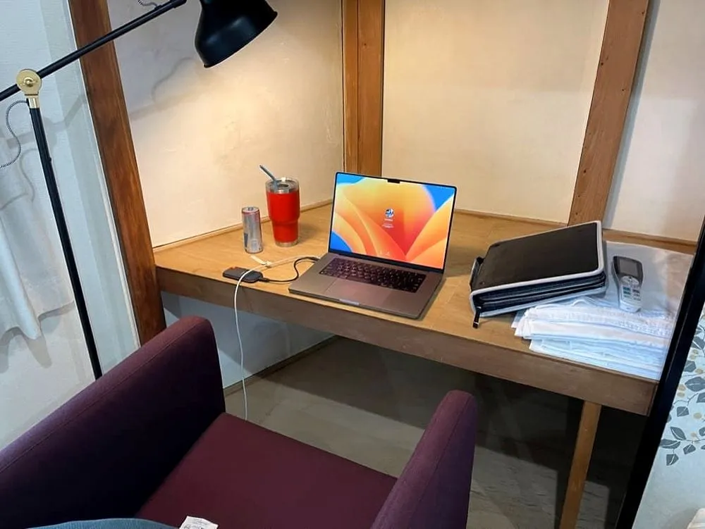 A wooden desk in a Japanese apartment in Osaka with an open MacBook, a red tumbler, an energy drink can, and a stack of papers under a floor lamp.