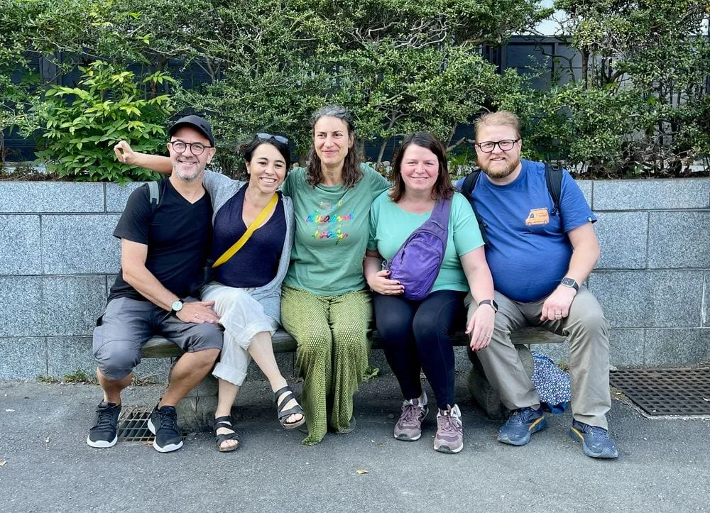 Adam and Lindsay sit on a low stone bench with three other adults in front of a green hedge, smiling at the camera on a street in Kawasaki, Japan.