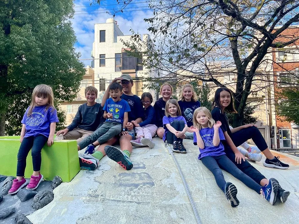 A group of worldschooling kids sitting together on a large concrete slide structure at a park in Taito, Tokyo, on a sunny October afternoon.