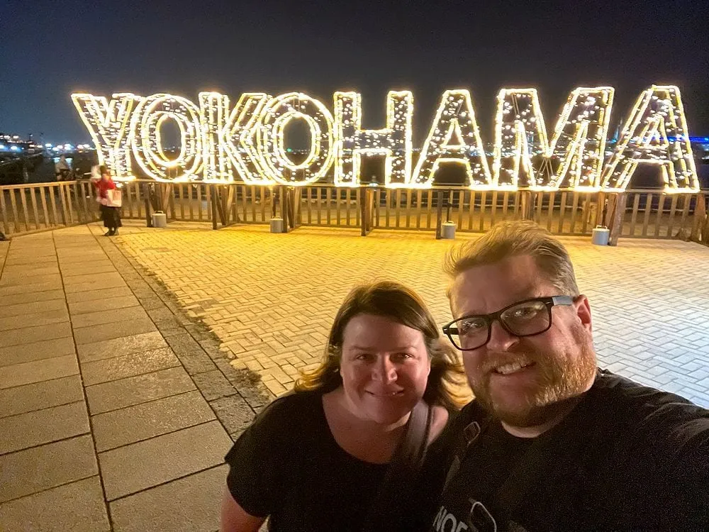 Adam and Lindsay take a selfie at night in front of a large illuminated YOKOHAMA sign made of string lights on the Shinko waterfront.