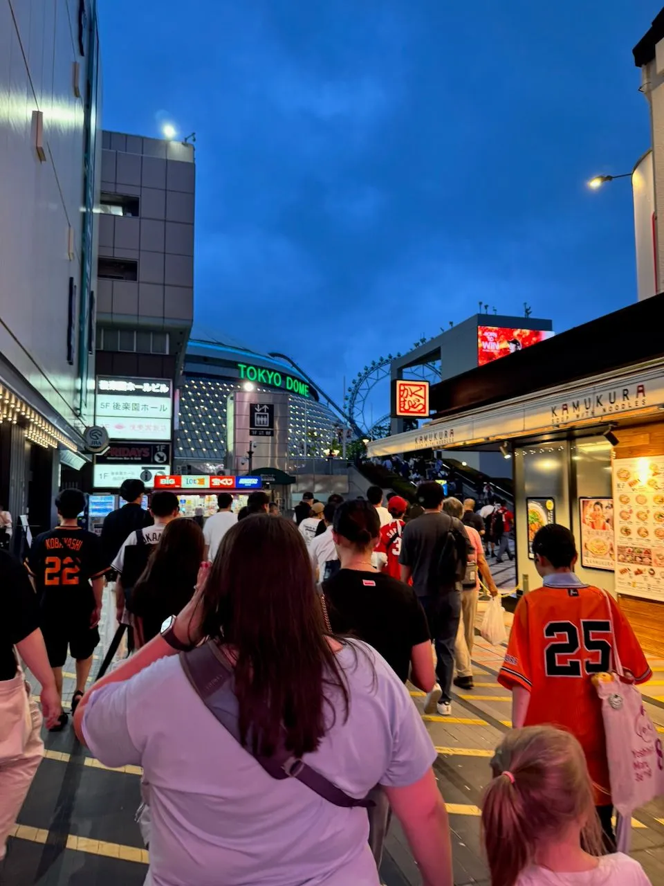 A crowd of people in baseball jerseys walking toward the illuminated Tokyo Dome sign at dusk, with a woman in a white shirt and a young blonde girl visible in the foreground.