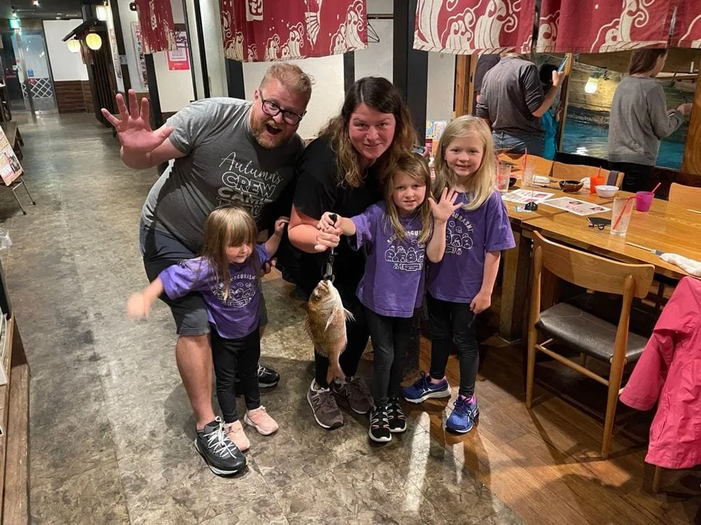 A family of five poses inside Zauo Fishing Restaurant in Namba, Osaka, with one of the girls holding up a whole raw fish on a hook, the group in matching purple shirts.