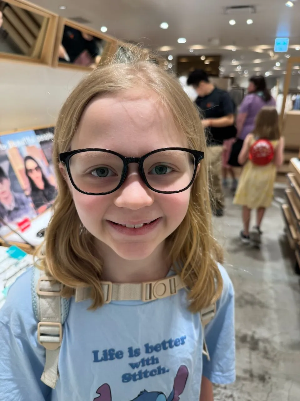 A young girl with blonde hair smiles at the camera wearing oversized black-framed glasses and a Stitch t-shirt inside a Japanese department store, surrounded by display cases of eyewear.