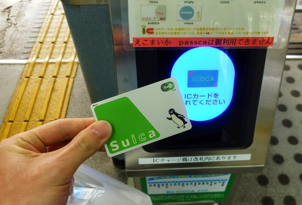 A Suica IC card with a penguin mascot design being tapped against a station gate reader, whose display also shows logos for ICOCA and other regional IC cards.